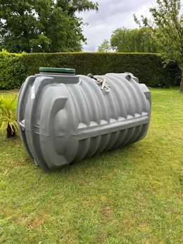 Réservoir cylindrique gris posé sur l'herbe dans un jardin, avec des haies et des arbres en arrière-plan.