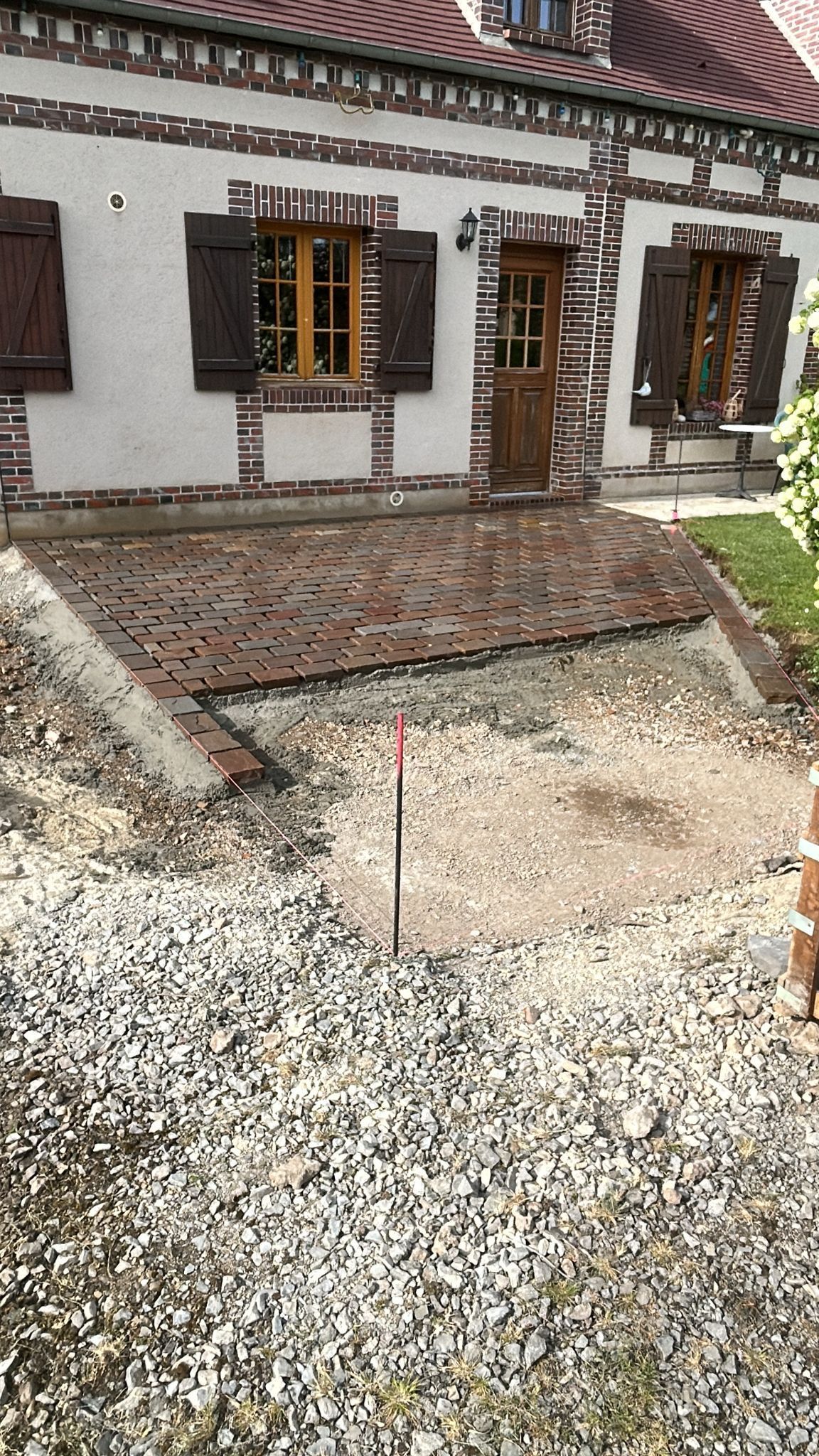 Extérieur de la maison avec un chemin de gravier et une petite terrasse pavée, murs blancs et fenêtres à cadre marron.