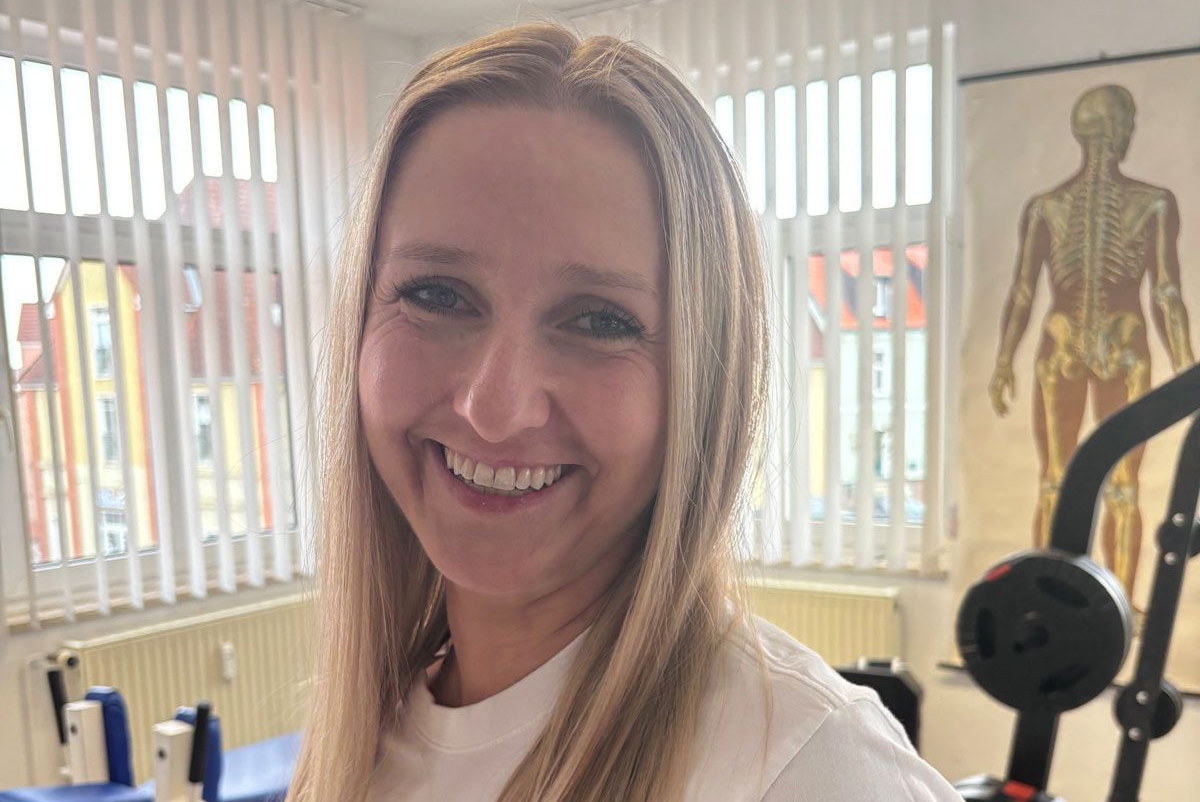 Eine lächelnde Person mit langen blonden Haaren in einem weißen Hemd, die in einer Physiotherapie- oder Fitnessstudio-Umgebung steht.