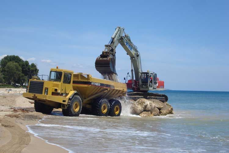Un camión volquete amarillo está siendo cargado con arena por una excavadora en una playa.