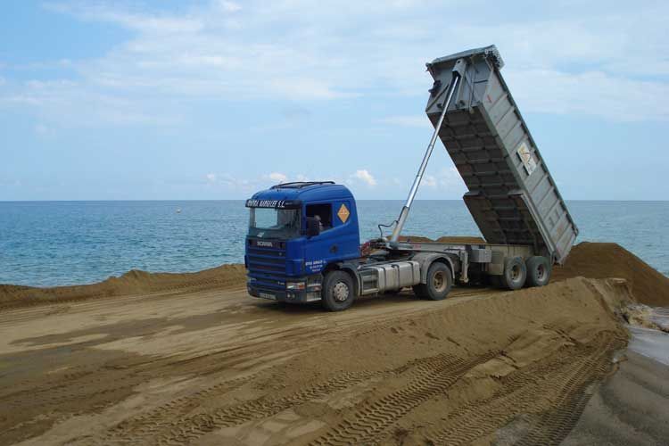 Un camión volquete azul circula por un camino de tierra cerca del océano.