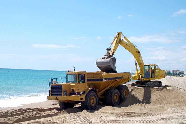 Una excavadora está cargando arena en un camión volquete en una playa.