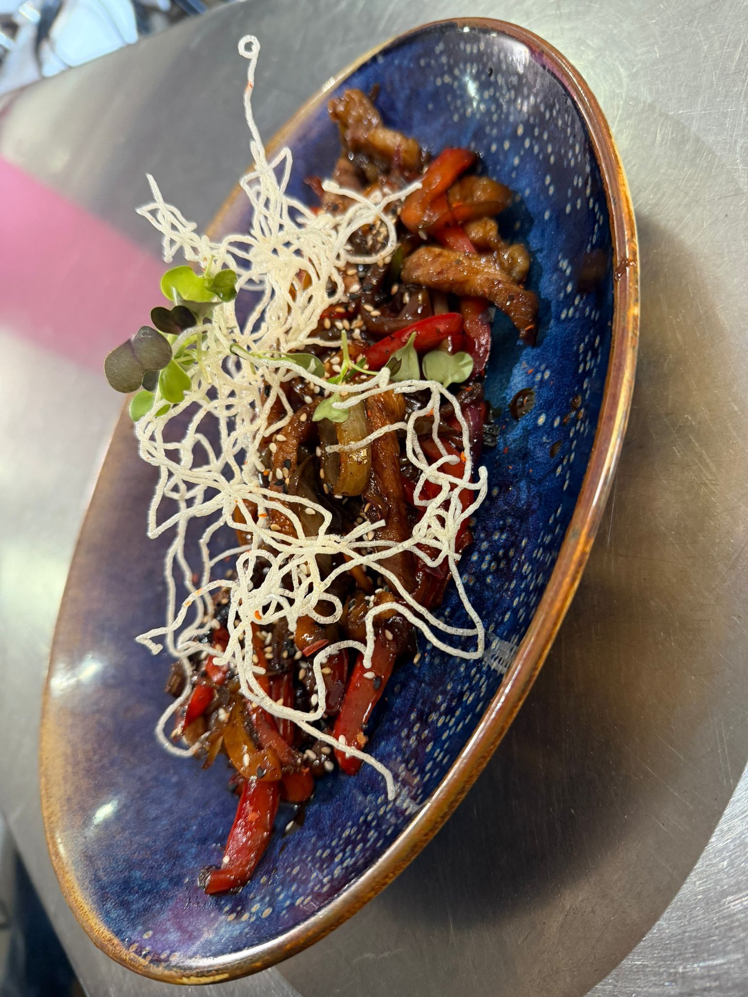 Plato azul con carne brillante en salsa, pimientos rojos y guarnición desmenuzada sobre una mesa.