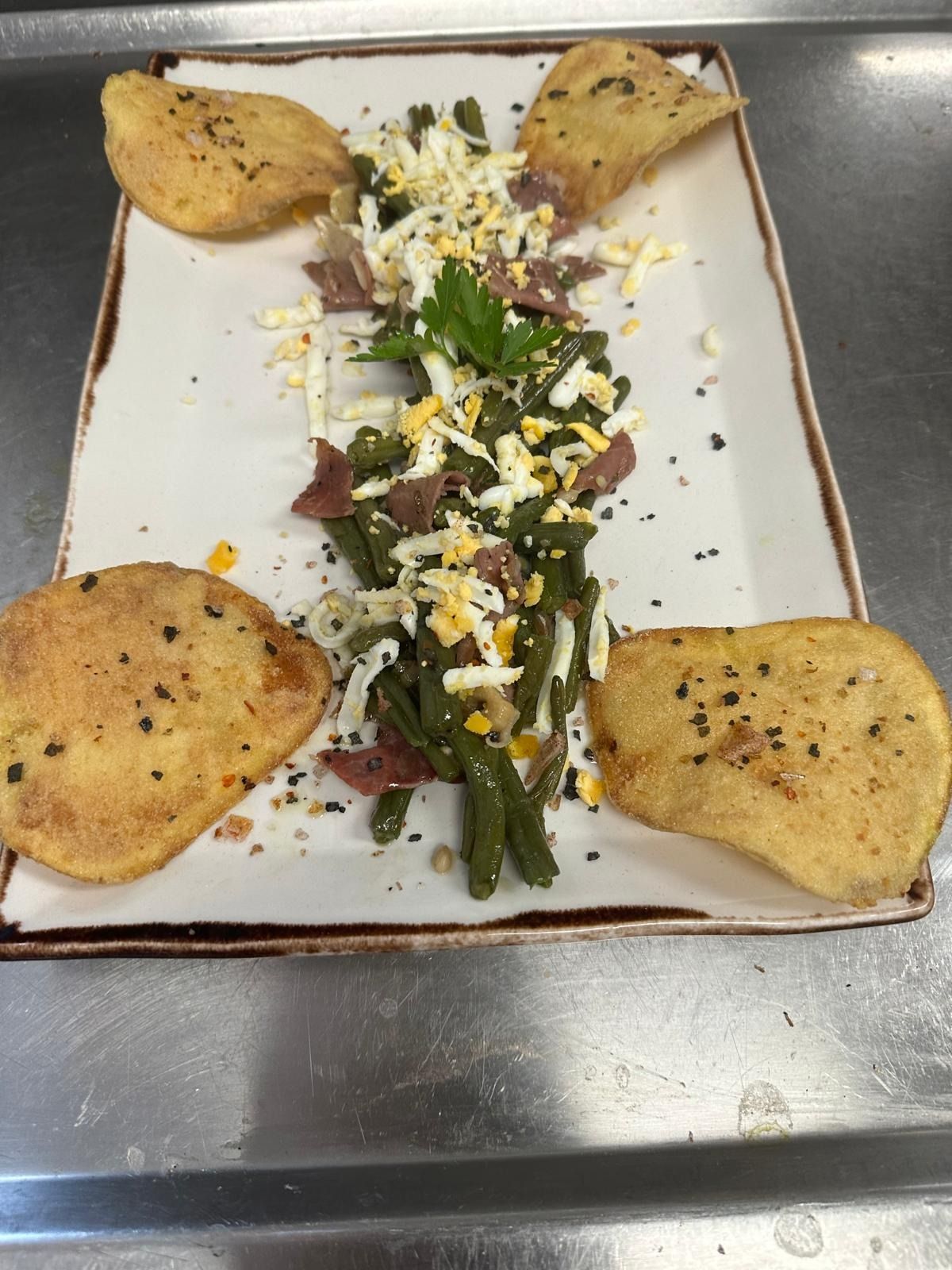 Plato rectangular blanco con una guarnición de verduras en el centro y cuatro gajos dorados y crujientes en las esquinas.