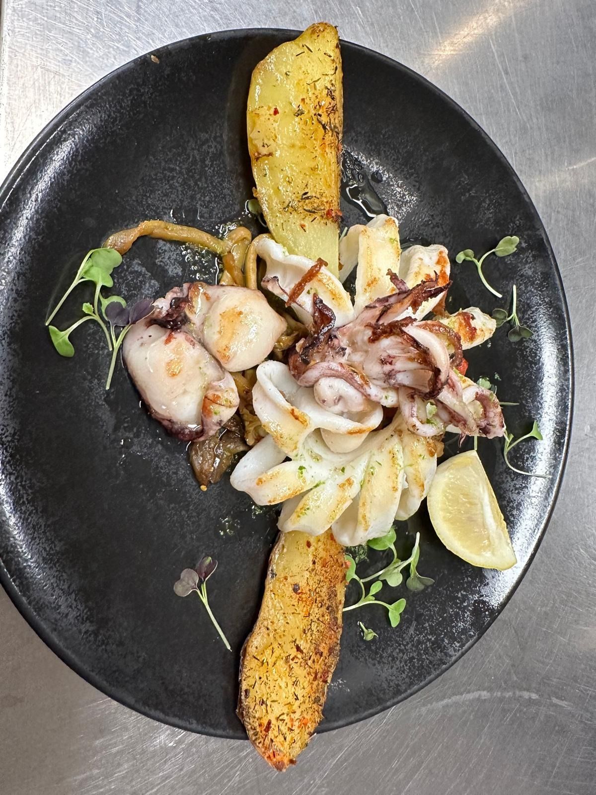 Plato de mariscos con pulpo a la parrilla, rodajas de limón y pan tostado en un plato negro.