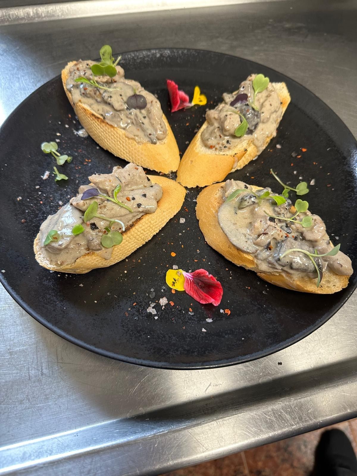 Cuatro rebanadas de bruschetta de champiñones sobre un plato negro, adornadas con microvegetales y flores comestibles.
