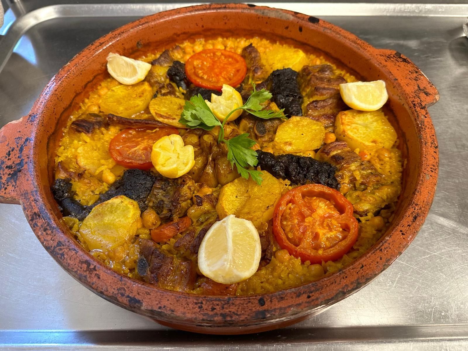 Plato de barro con arroz y carne horneados, adornado con rodajas de limón y hierbas, sobre una superficie metálica.