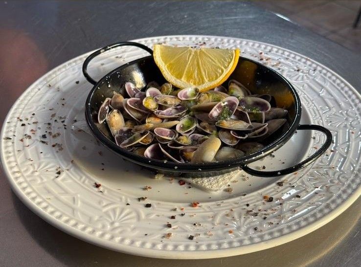 Sartén negra con almejas en caldo sobre un plato blanco, adornada con una rodaja de limón.