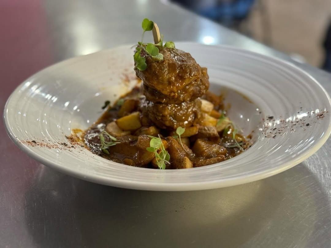Bol blanco con comida gourmet en salsa, cubierto con hierbas, sobre una mesa de acero inoxidable.