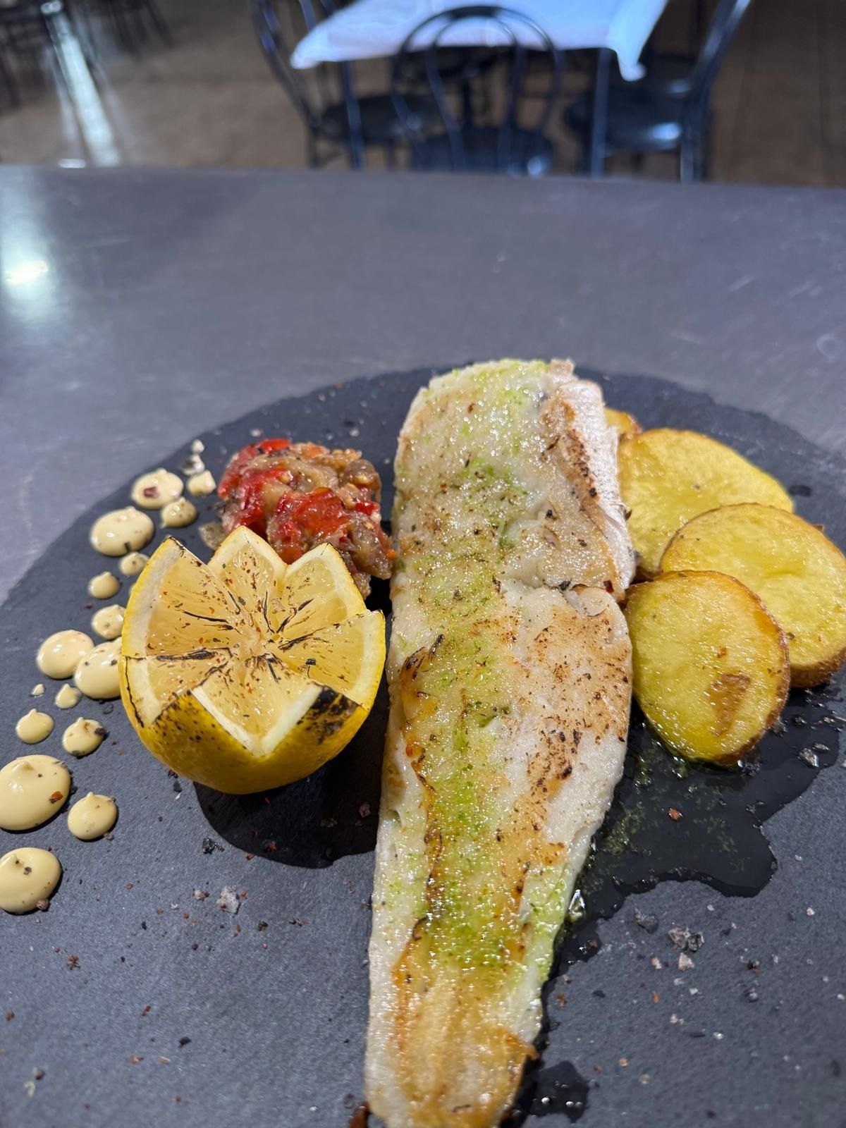 Filete de pescado a la parrilla con limón, patatas asadas y salsa en un plato oscuro.