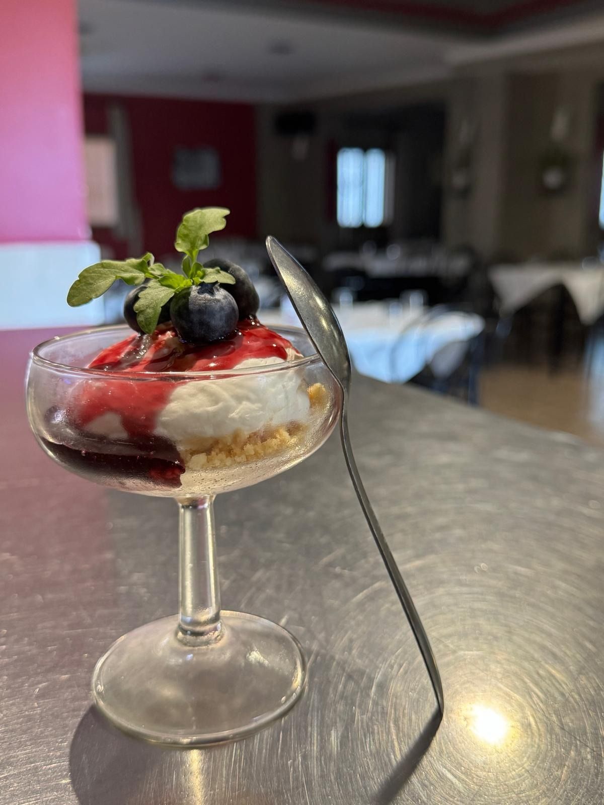Parfait de postre con frutos rojos y menta en una copa con tallo, junto a una cuchara sobre una mesa.
