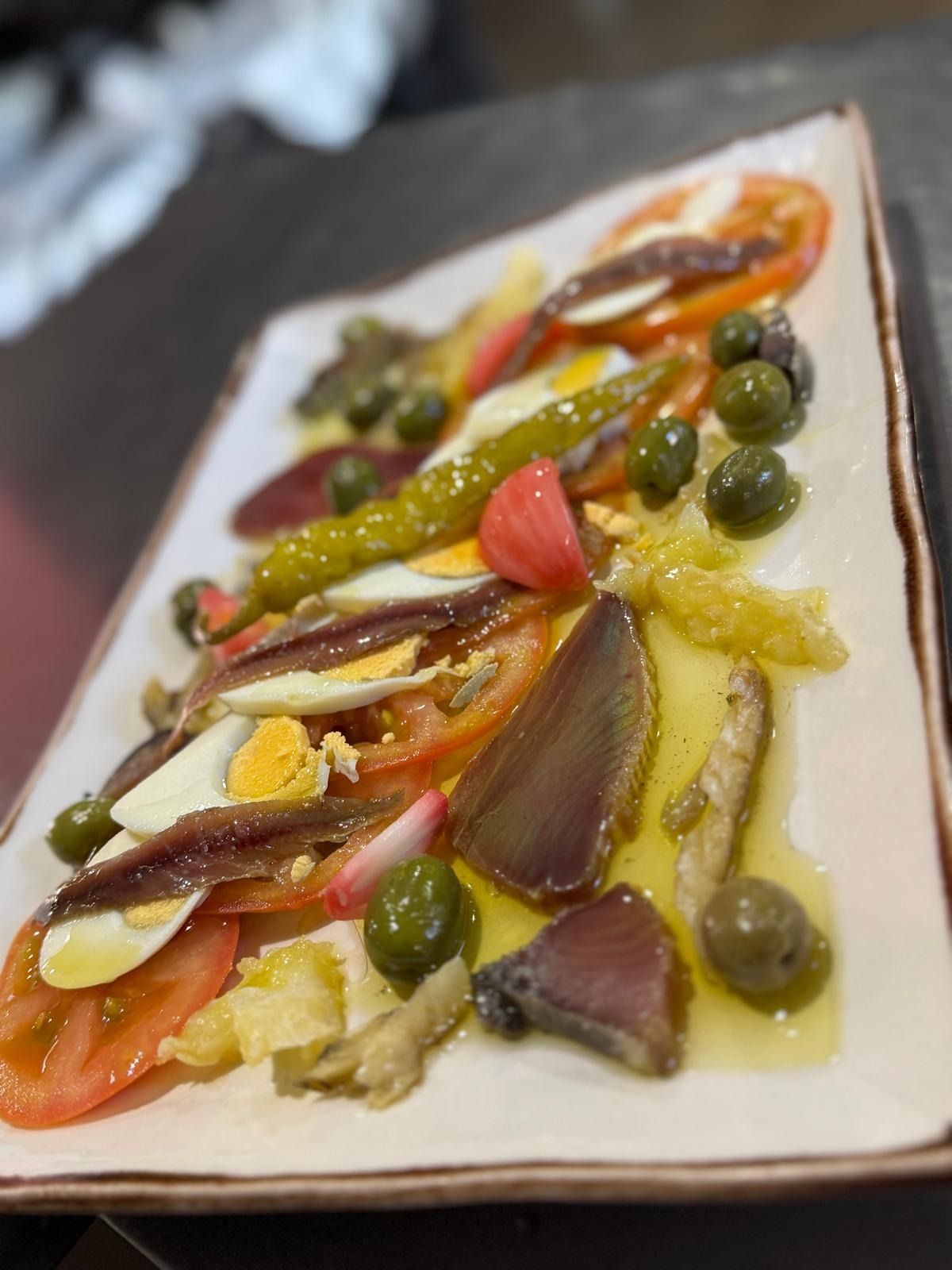 Plato gourmet presentado con coloridas verduras, aceitunas y salsas en un plato blanco rectangular.
