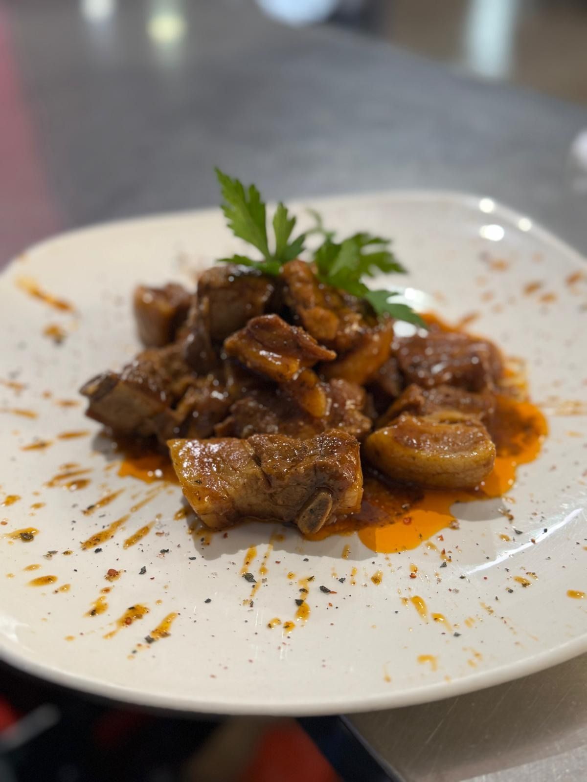 Carne estofada emplatada en salsa marrón con hierbas sobre un plato blanco, con salpicaduras de aceite de naranja.