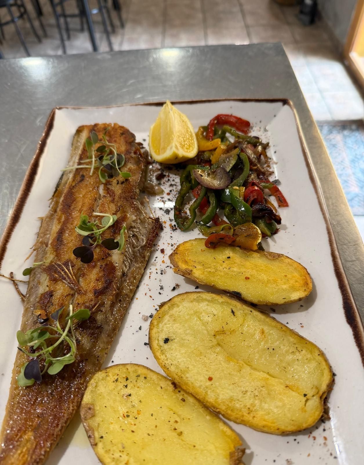Pescado a la parrilla emplatado con una rodaja de limón, verduras salteadas y dos mitades de patata asada en un plato blanco.