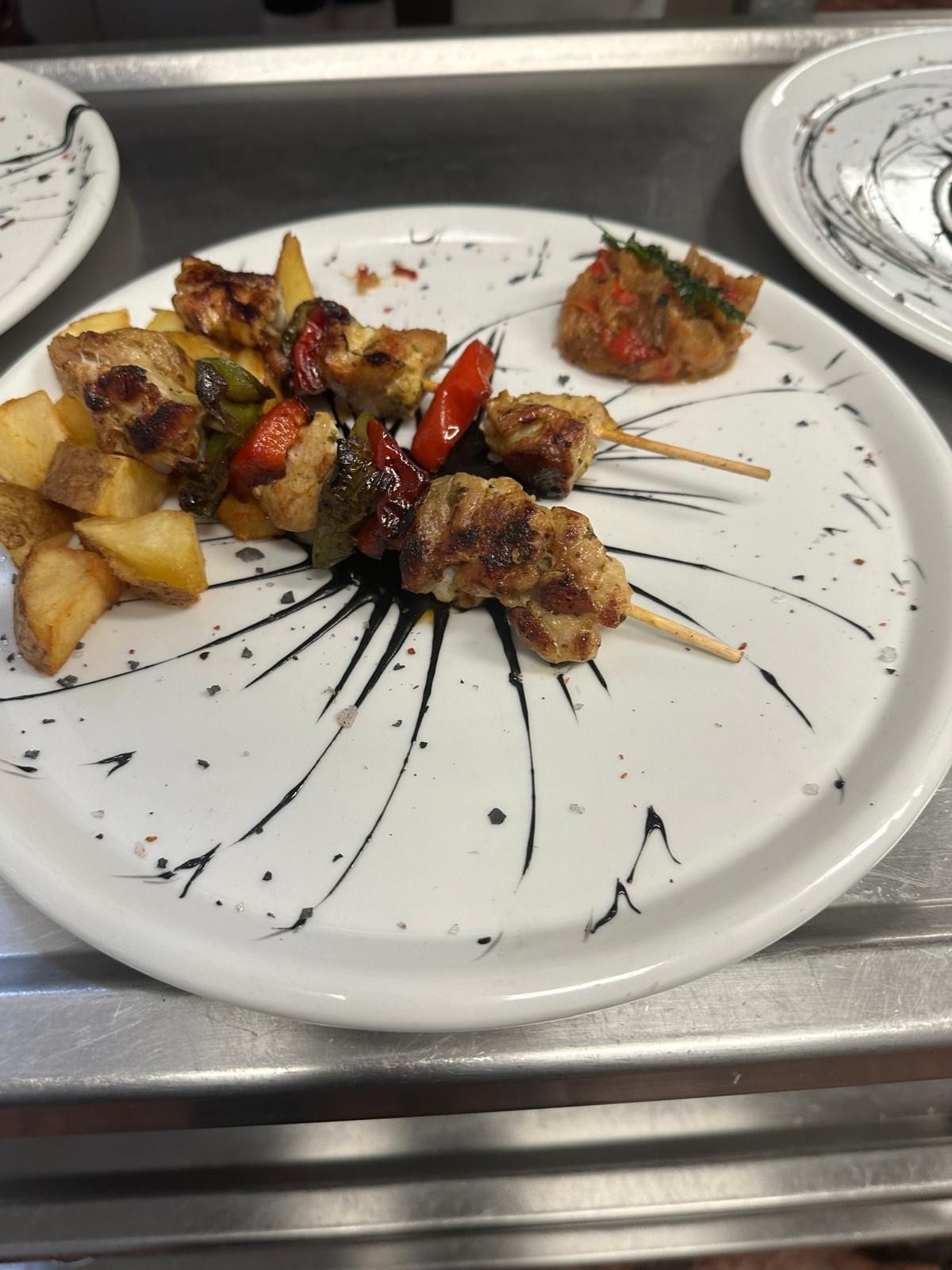 Brochetas de carne y verduras a la parrilla servidas en un plato blanco con un chorrito de salsa negra y una guarnición