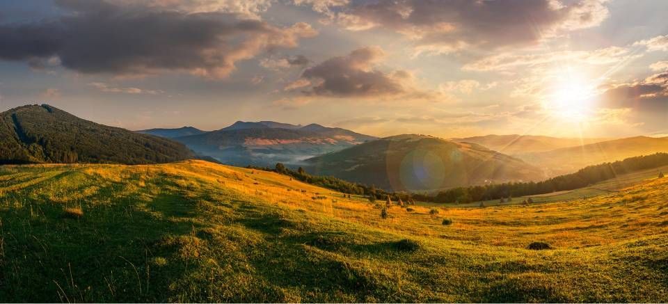 Al tramonto, la luce dorata del sole illumina un prato erboso, con dolci catene montuose sullo sfondo, sotto un cielo nuvoloso.