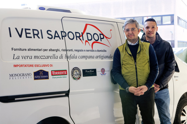 Olio d'oliva dorato viene versato da una bottiglia di vetro in una piccola ciotola, circondata da olive fresche e foglie su una superficie di legno.