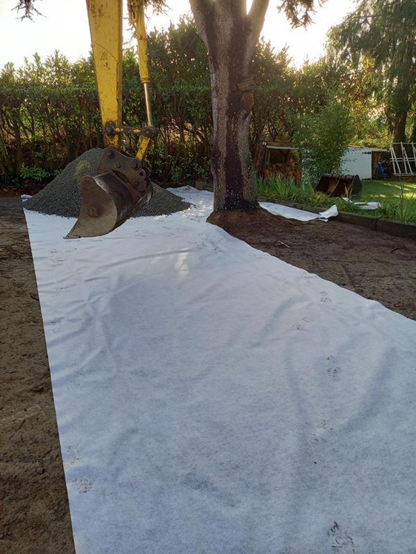 Tissu blanc disposé dans une cour près d'un arbre, avec une pelle rétrocaveuse et un tas de gravier en arrière-plan.