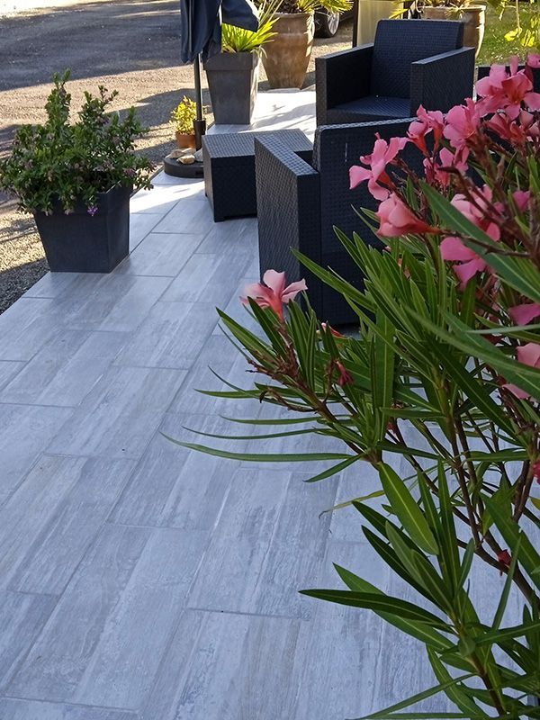 Terrasse avec carrelage imitation bois gris, mobilier noir, fleurs roses et plantes en pot. Cadre extérieur ensoleillé.
