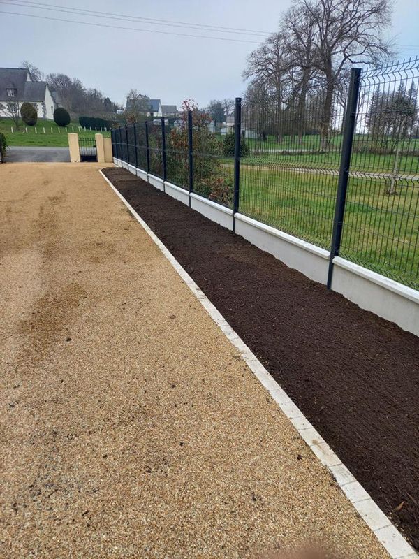 Une allée avec du gravier à gauche et une bande de terre foncée à droite, bordée d'un mur blanc et d'une clôture noire.