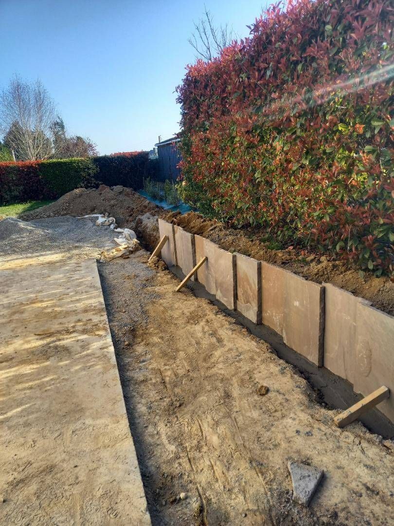 Un mur de soutènement en bois en cours de construction le long d'un chemin de terre, à côté d'une haie sous un ciel bleu.