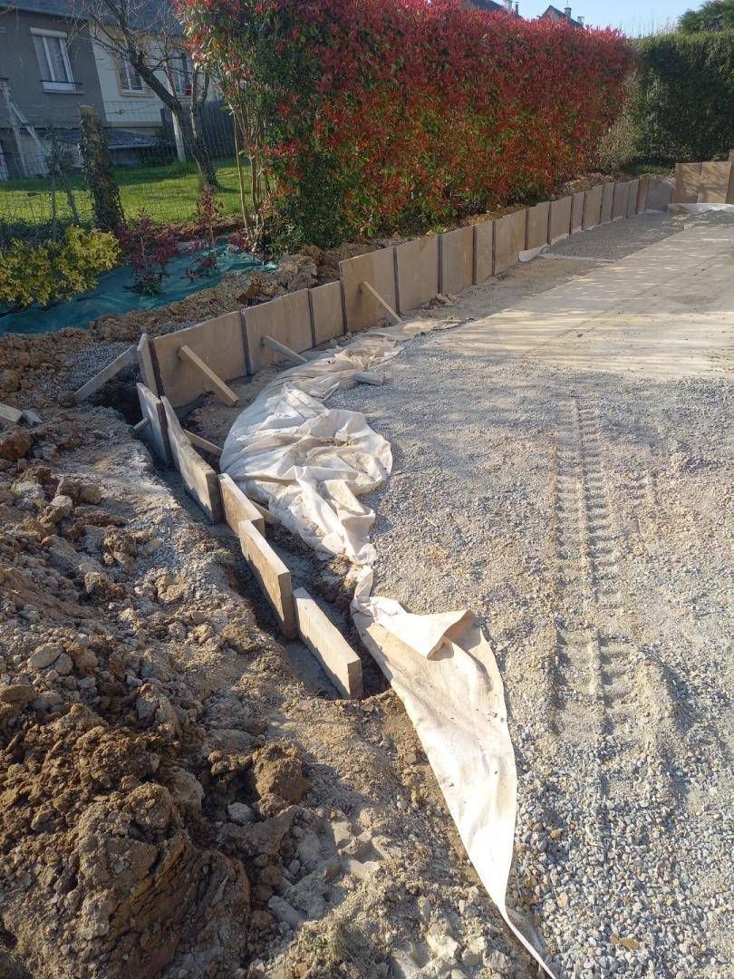 Chantier de construction avec coffrages en bois pour un mur de béton le long d'une allée de gravier.