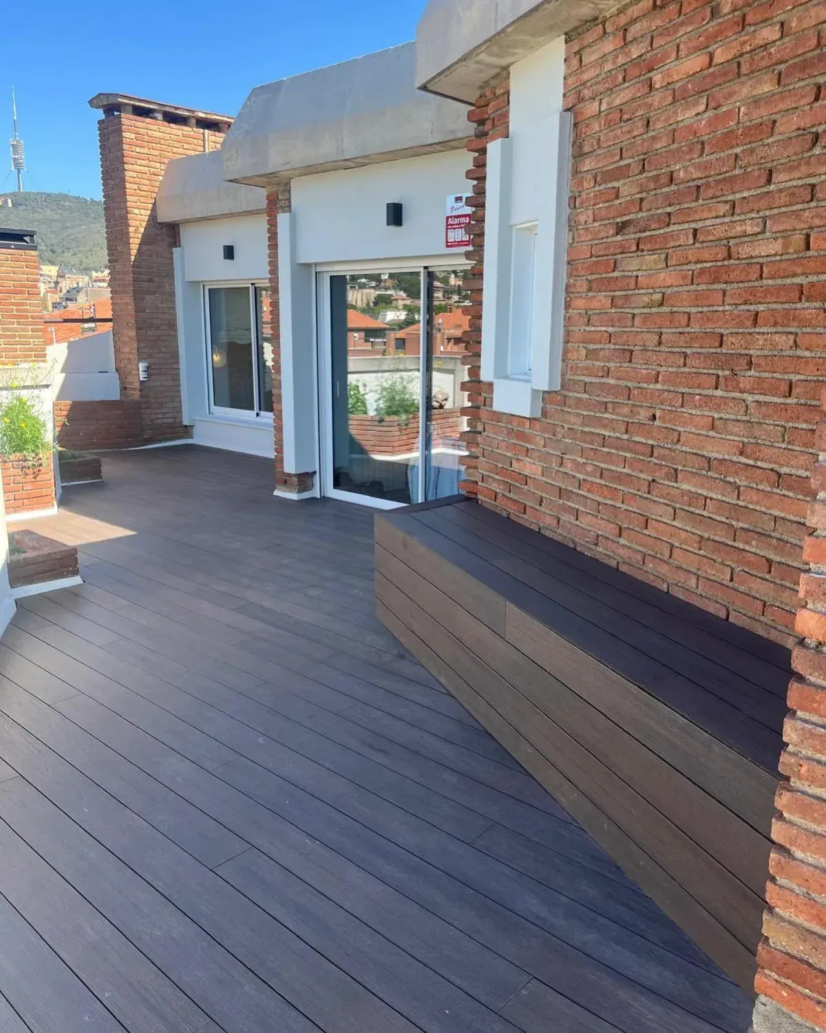 Terraza en la azotea con tarima compuesta, paredes de ladrillo, un banco y puerta de vidrio.