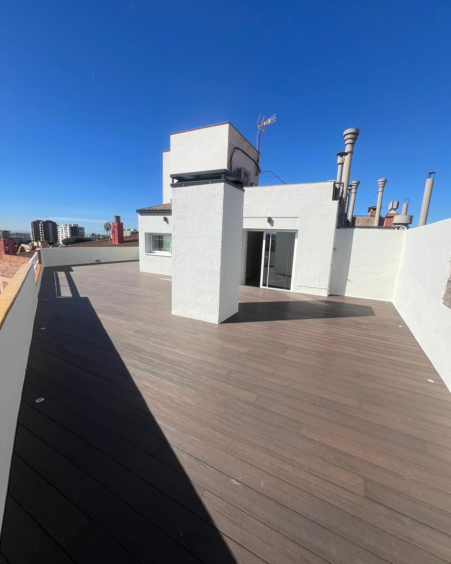 Terraza en la azotea con suelo de madera oscura, paredes blancas y un cielo azul claro.