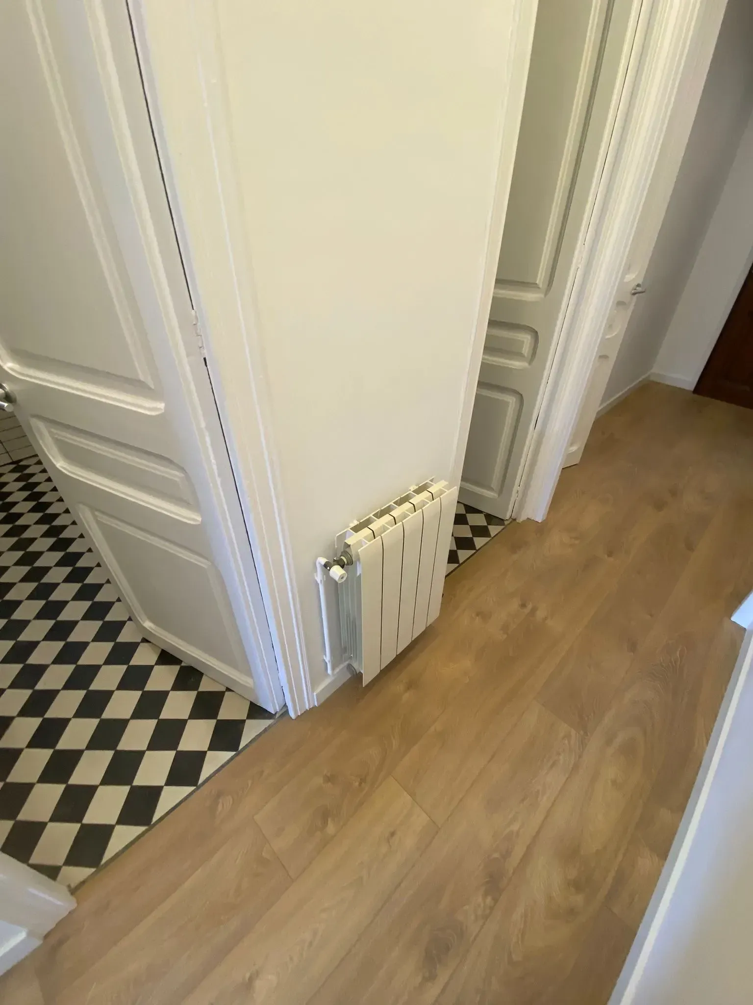 Pasillo blanco con suelo de madera y radiador en la pared. Suelo de baldosas blancas y negras visible a la izquierda.