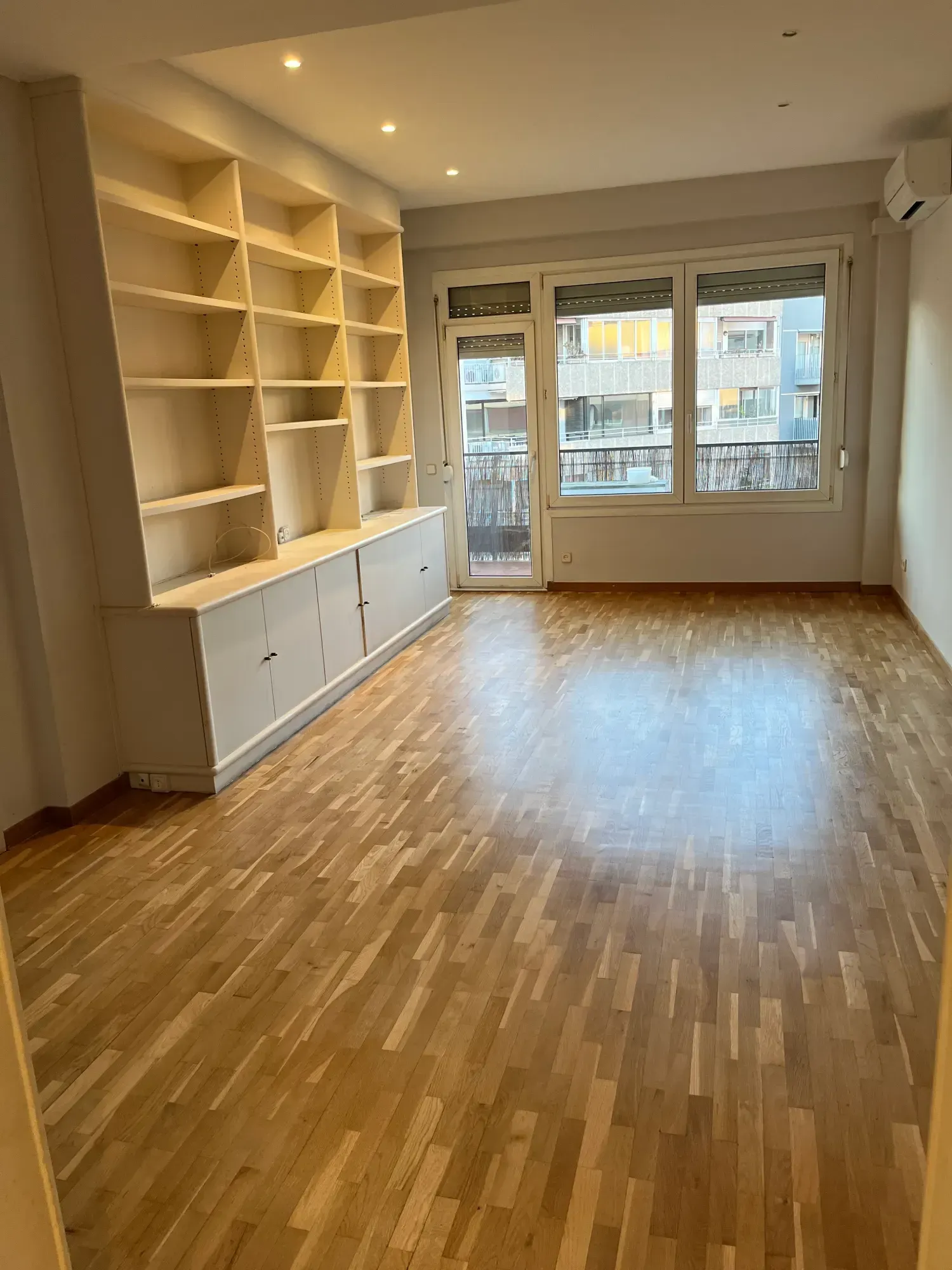 Habitación vacía con pisos de madera, estanterías empotradas y una puerta de balcón que da a una calle de la ciudad.