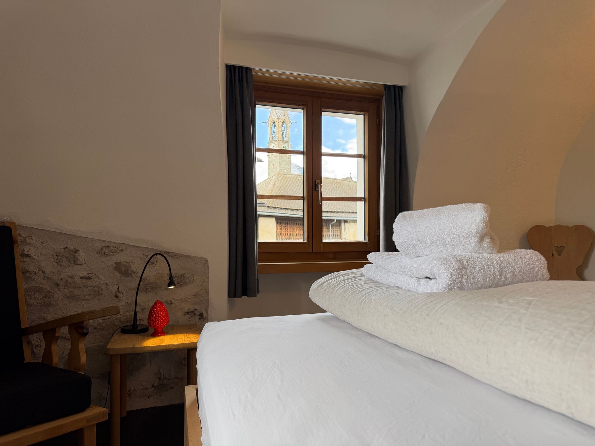 Helles Schlafzimmer mit Holzbett, weisser Bettwäsche und zwei Fenstern mit dunklen Vorhängen.