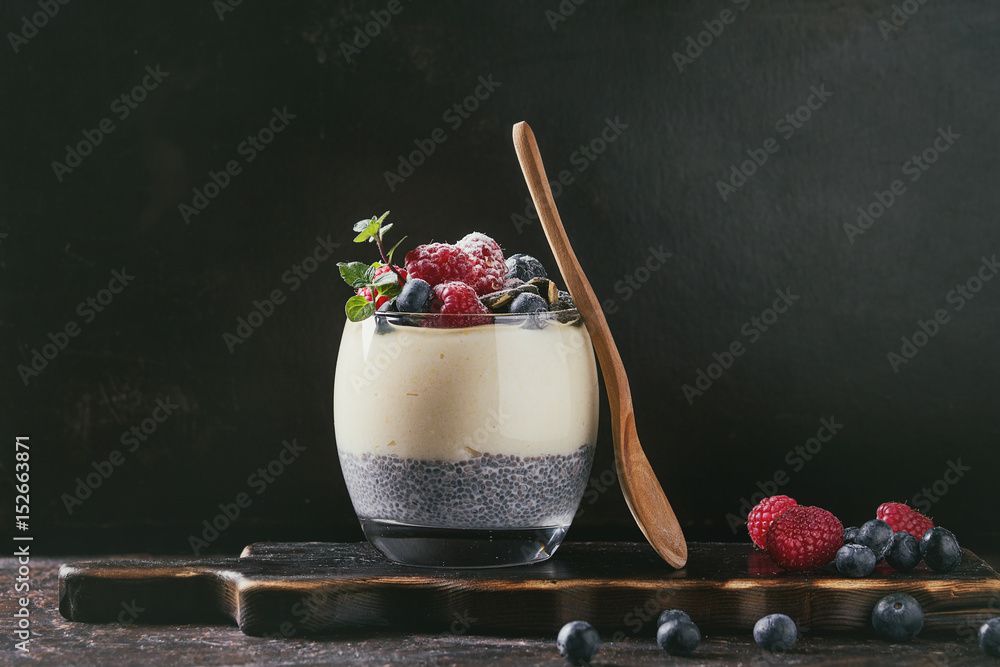 Ein Glas mit geschichtetem Dessert aus Blaubeeren, Himbeeren und einem Holzlöffel auf einem Holzbrett.