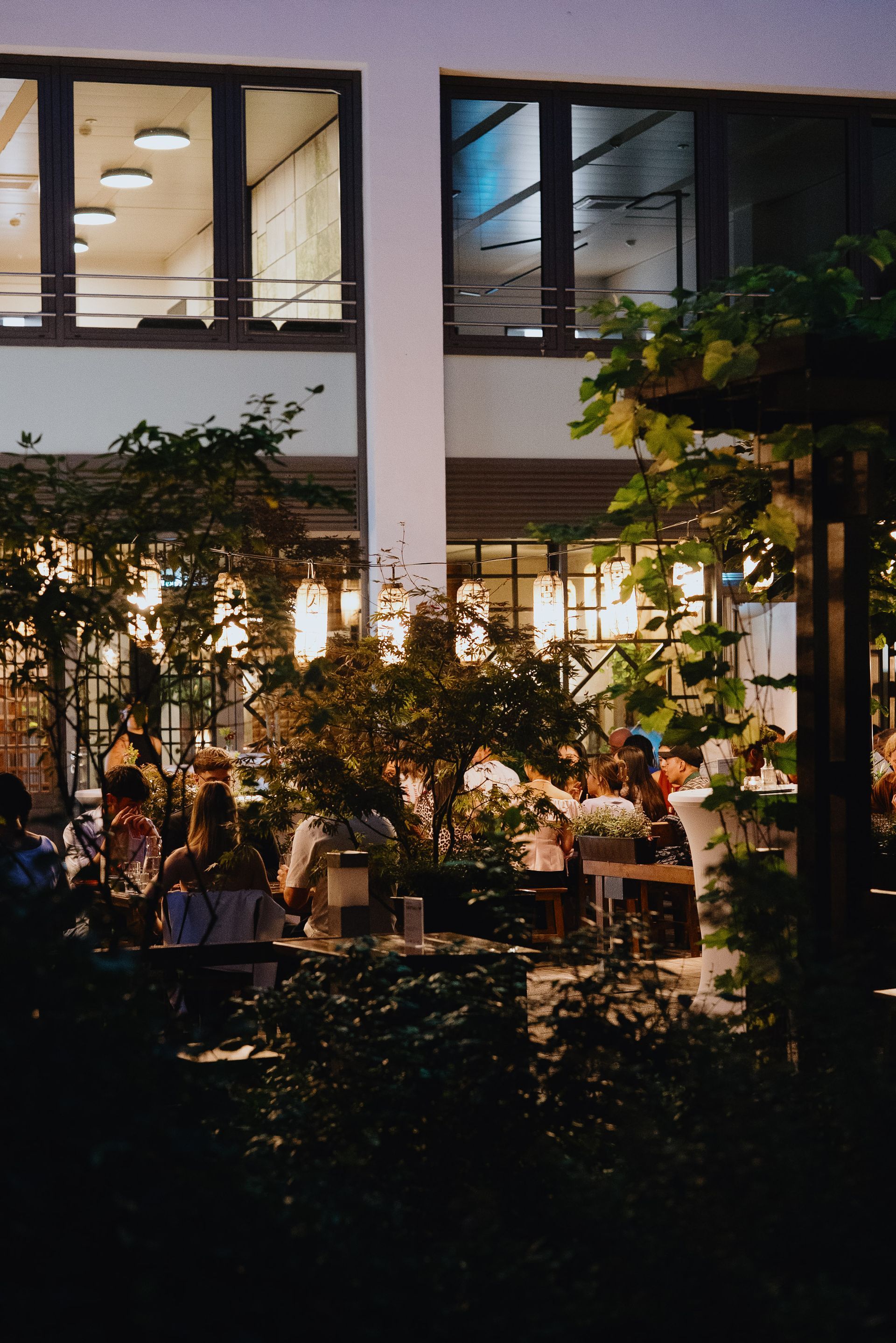 Essbereich im Freien mit beleuchteten Papierlaternen. Gäste sitzen an Tischen, umgeben von Grün.