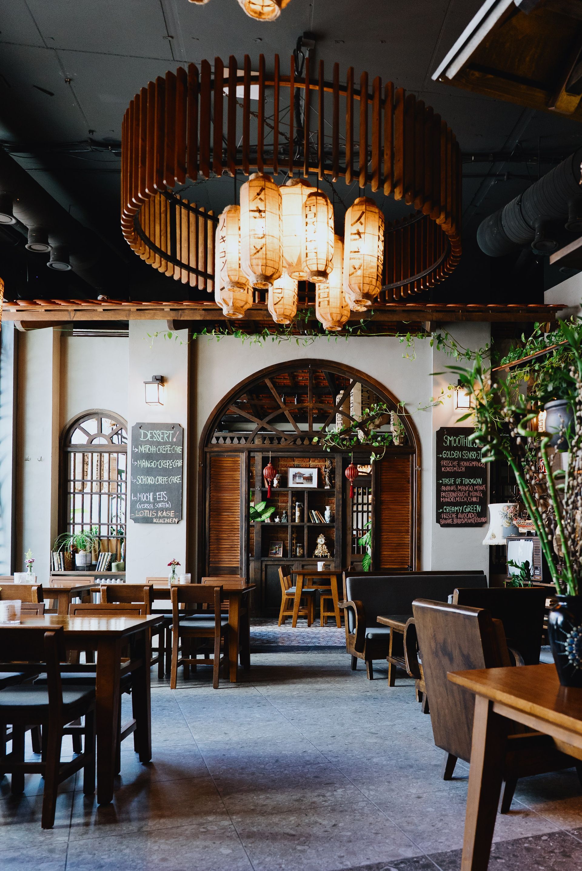 Restauranteingang mit gewölbter Holztür, Tischen und Pflanzen unter einem hölzernen Rankgitter.