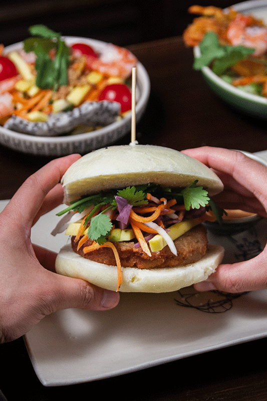 Hände halten ein mit Füllung und Grünzeug belegtes Bao-Bun-Sandwich neben Salatschüsseln.