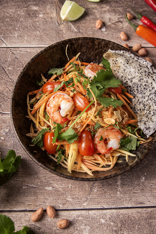 Garnelen-Papaya-Salat in einer dunklen Schüssel mit Erdnüssen, Tomaten und einem knusprigen Cracker auf einer Holzoberfläche.