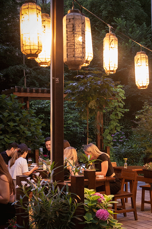 Essbereich im Freien mit beleuchteten Papierlaternen, Tischen, Stühlen und essenden Menschen.