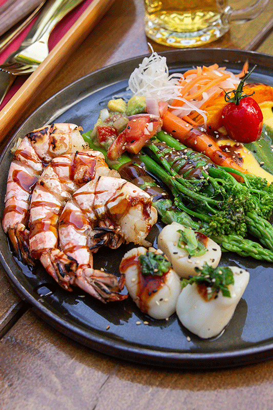 Gegrillte Garnelen und Jakobsmuscheln mit Gemüse auf einem Teller, daneben ein Glas Bier und Besteck.