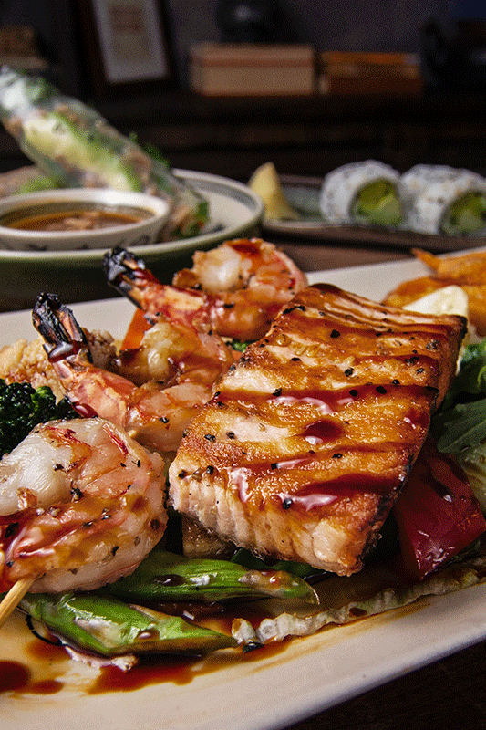 Gegrillter Lachs, Garnelenspieße und Gemüse mit Dip-Sauce auf einem Teller, Sushi im Hintergrund.