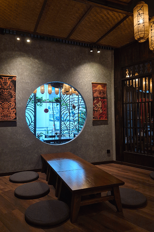 Zimmer im asiatischen Stil mit einem runden Fenster zum Innenhof. Niedriger Tisch mit Kissen, Holzelemente und Hängelaternen.