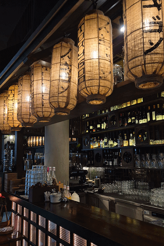 Interieur einer Bar, beleuchtet von Papierlaternen. Im Vordergrund befindet sich eine hölzerne Bar, im Hintergrund Regale mit Spirituosen.