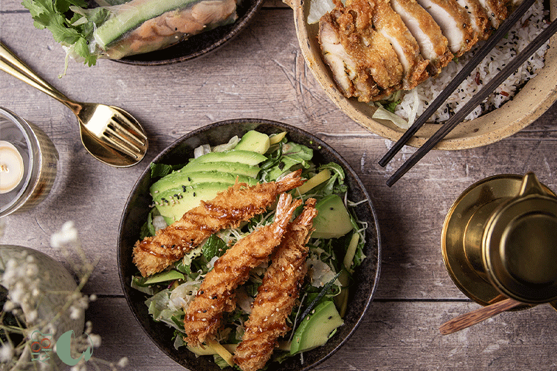 Asiatisch inspirierte Gerichte: frittierte Garnelen und Avocadosalat, Frühlingsrollen und Hühnchen mit Reis, alles auf einem Holztisch serviert.