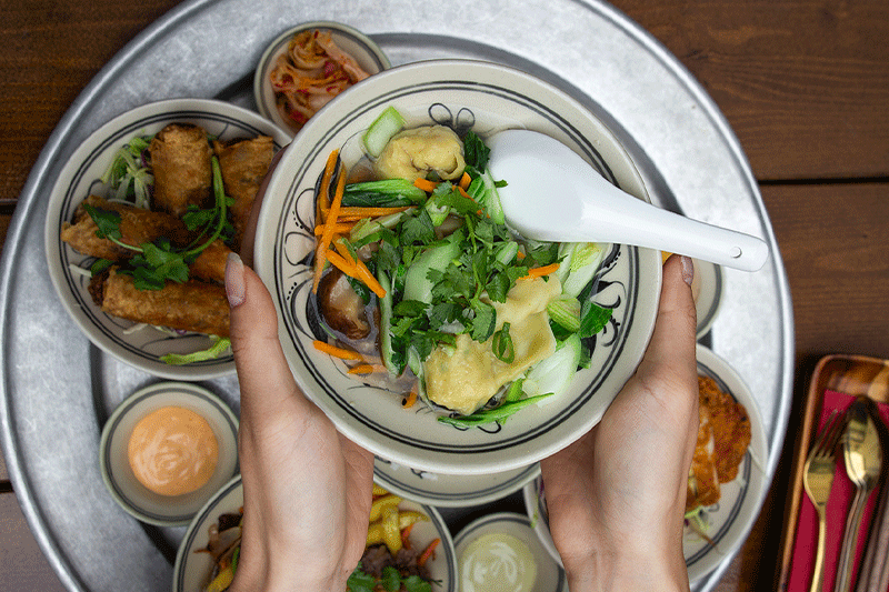 Hände halten eine Schüssel mit vietnamesischer Suppe, umgeben von verschiedenen Beilagen auf einem silbernen Tablett.