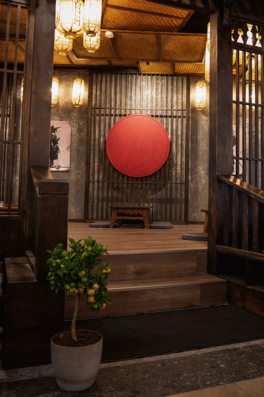 Restauranteingang im japanischen Stil: rote Kreiskunst, Holzplattform, Topfpflanze, Papierlaternen, dunkles Holz.
