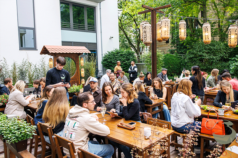 Restaurant im Freien mit Gästen an Tischen, Dekoration im japanischen Stil, hängende Laternen.