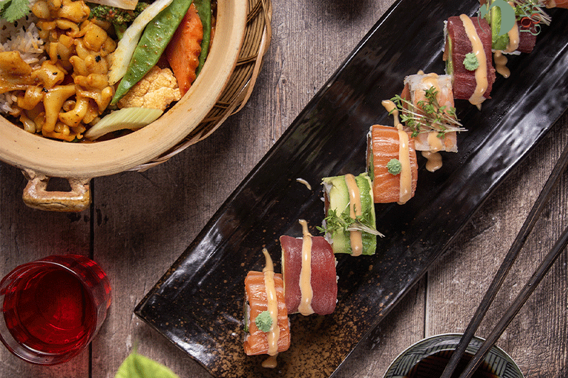 Sushi-Rollen mit Sauce auf einer schwarzen Platte, dazu eine Schüssel mit Essen und ein Glas rote Flüssigkeit.