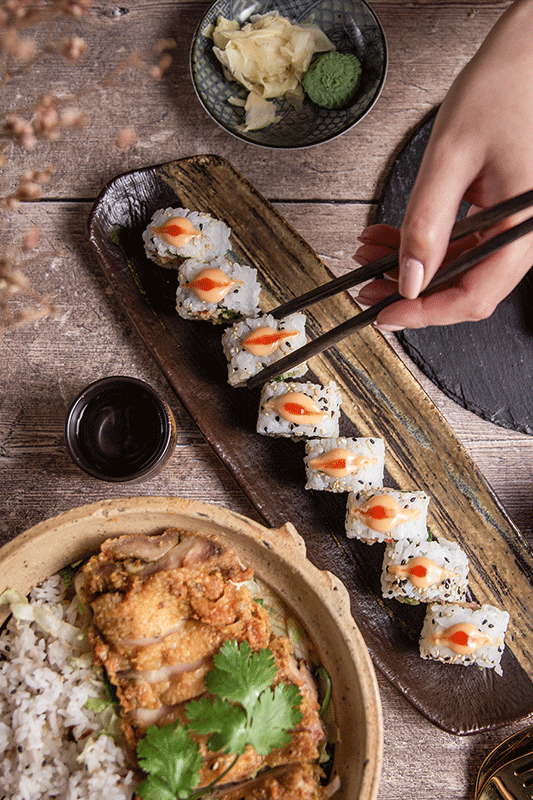 Sushi und gebratenes Hähnchen auf einem Holztisch, Essstäbchen greifen nach einer Sushi-Rolle.