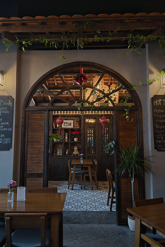 Außenansicht eines Restaurants mit Holztischen und einem großen Bogeneingang. Der Innenraum mit Dekoration und Sitzgelegenheiten ist sichtbar.