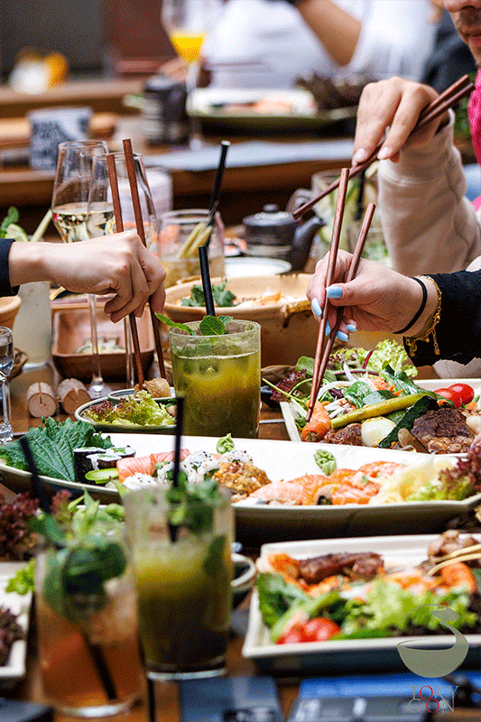 Menschen, die an einem Restauranttisch mit Speisen, Getränken und Grünzeug Stäbchen benutzen.