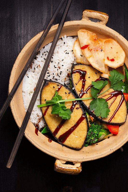 Eine Schüssel mit Reis, Tofu und Gemüse, dazu Essstäbchen. Mit Soße beträufelt und mit Kräutern garniert.